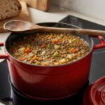 Hrnec plný čočkové polévky se zeleninou na sklokeramické desce.