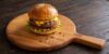 Hotový, bohatě vrstvený domácí hovězí burger v sezamové bulce s rozteklým sýrem, karamelizovanou cibulí, salátem a rajčetem, servírovaný na masivním dřevěném prkénku.