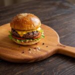 Hotový, bohatě vrstvený domácí hovězí burger v sezamové bulce s rozteklým sýrem, karamelizovanou cibulí, salátem a rajčetem, servírovaný na masivním dřevěném prkénku.
