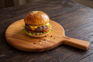 Hotový, bohatě vrstvený domácí hovězí burger v sezamové bulce s rozteklým sýrem, karamelizovanou cibulí, salátem a rajčetem, servírovaný na masivním dřevěném prkénku.