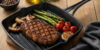 Grilovaný hovězí steak s chřestem, žampiony a rajčaty na černé grilovací pánvi na dřevěném stolu.
