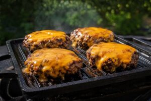 Detailní záběr na čtyři šťavnaté hovězí burgery s rozteklým sýrem čedar, které se opékají na černé litinové grilovací pánvi venku na grilu.